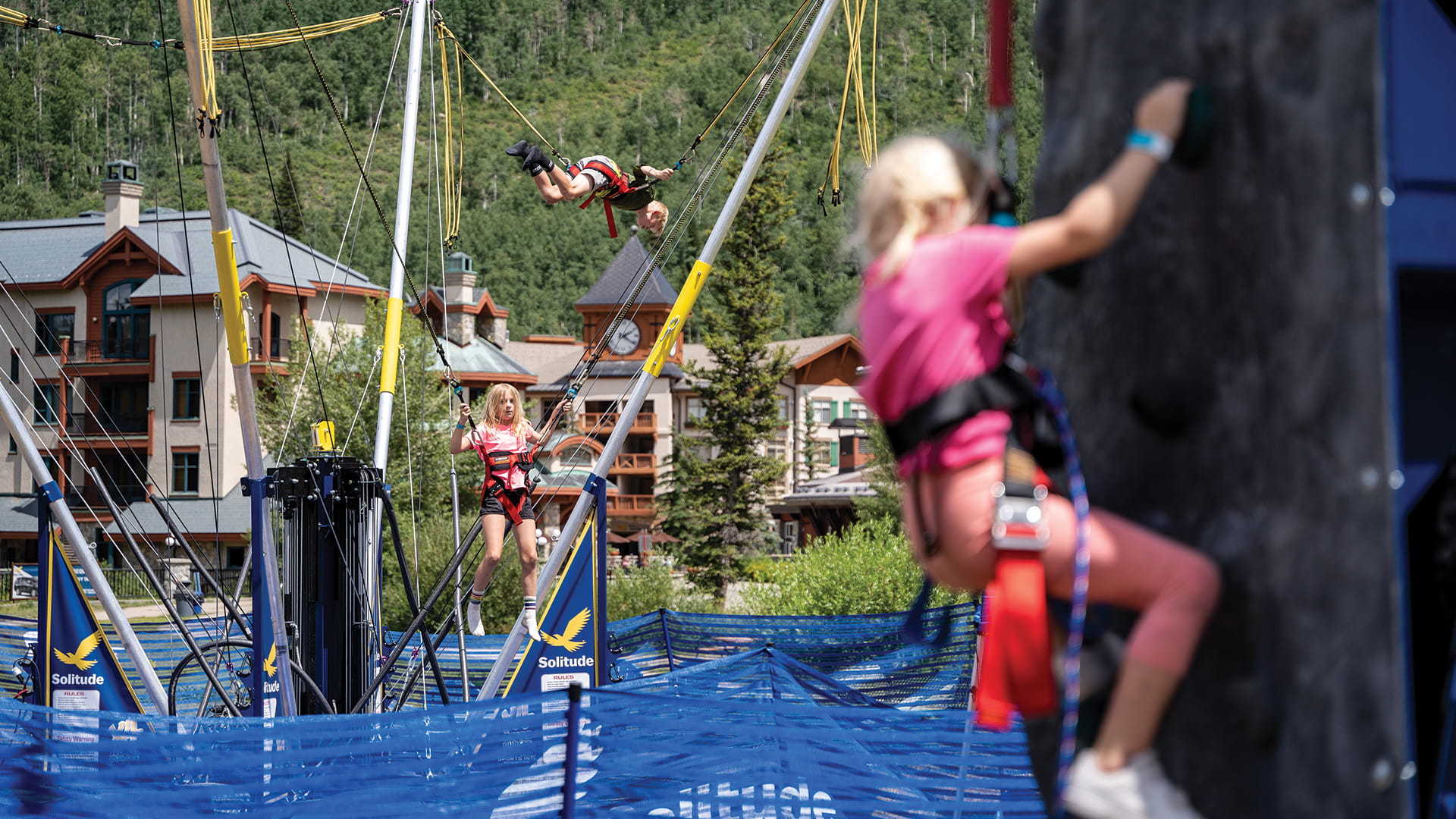 Kid climbing on the climbing wall with more kids on the bungee trampolines at Solitude Adventure Park 