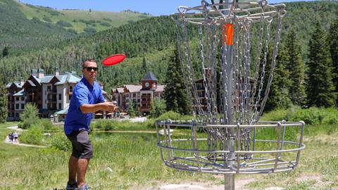 A disc golfer throwing a disc at the basket with the village in the background